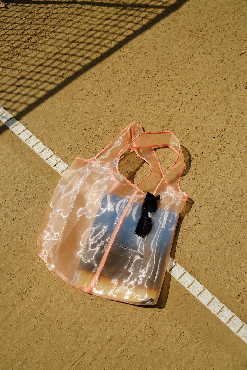 May include: A sheer pink mesh tote bag with a magazine and sunglasses inside. The bag is lying on a brown surface with a white line.
