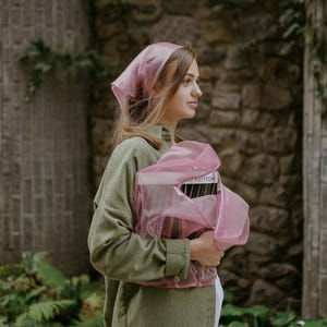 May include: A woman wearing a pink headscarf and a green shirt holds a pink bag with the text "LOUIS VUITTON" printed on it.
