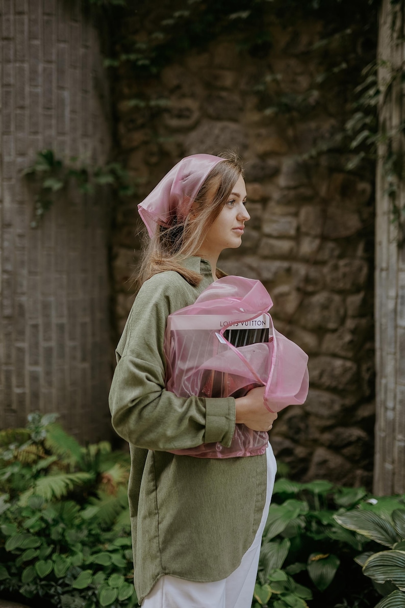 May include: A woman wearing a pink headscarf and a green shirt holds a pink bag with the text "LOUIS VUITTON" printed on it.