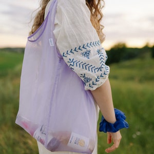 May include: A woman wearing a white dress with blue embroidery carries a purple mesh tote bag with two glass bottles inside. The bottles have labels that read "Kombucha" and "Est. 2021".