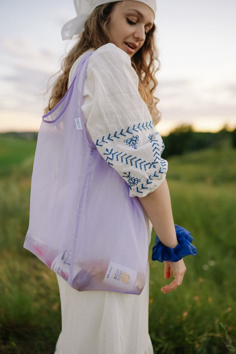 May include: A woman wearing a white dress with blue embroidery carries a purple mesh tote bag with two glass bottles inside. The bottles have labels that read "Kombucha" and "Est. 2021".
