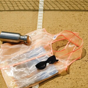 May include: A pink mesh tote bag with a white label that says "Tote". The bag is lying on a brown surface with a white line running through the middle. A silver water bottle and sunglasses are inside the bag.