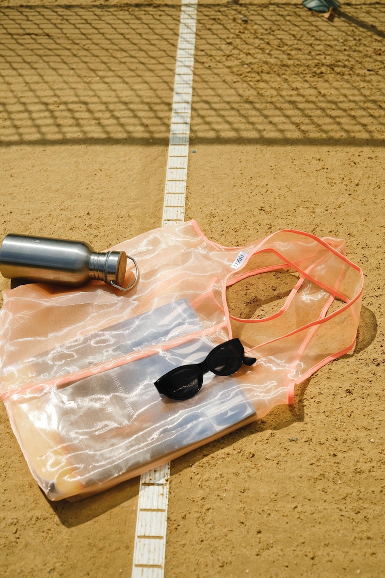 May include: A pink mesh tote bag with a white label that says "Tote". The bag is lying on a brown surface with a white line running through the middle. A silver water bottle and sunglasses are inside the bag.