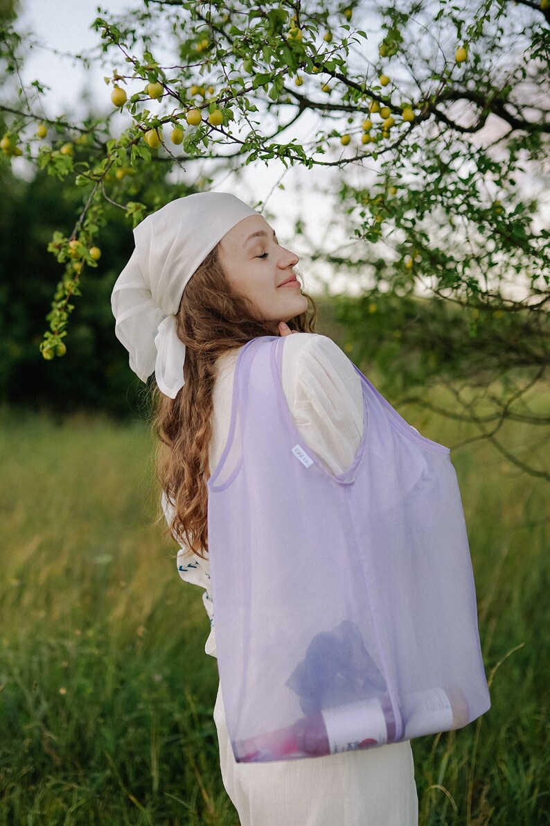 May include: A woman wearing a white headscarf and a white dress is holding a lavender mesh tote bag. The bag is filled with items, and the word "LOVE" is visible on the bag.