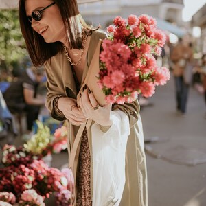 May include: A woman in a beige trench coat holds a bouquet of pink flowers in a brown paper bag. She is wearing sunglasses and a gold necklace.