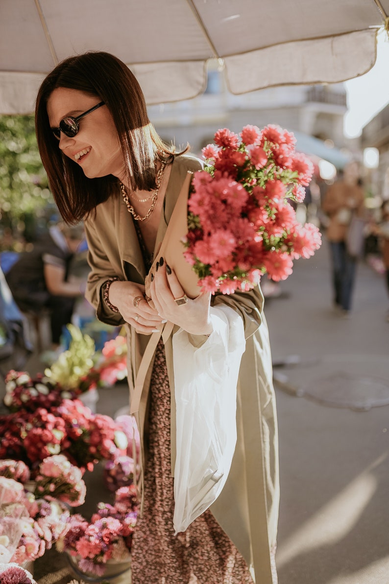 May include: A woman in a beige trench coat holds a bouquet of pink flowers in a brown paper bag. She is wearing sunglasses and a gold necklace.