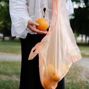 May include: A person holding a peach-colored mesh bag with oranges inside. The bag has a label that says "FREI."
