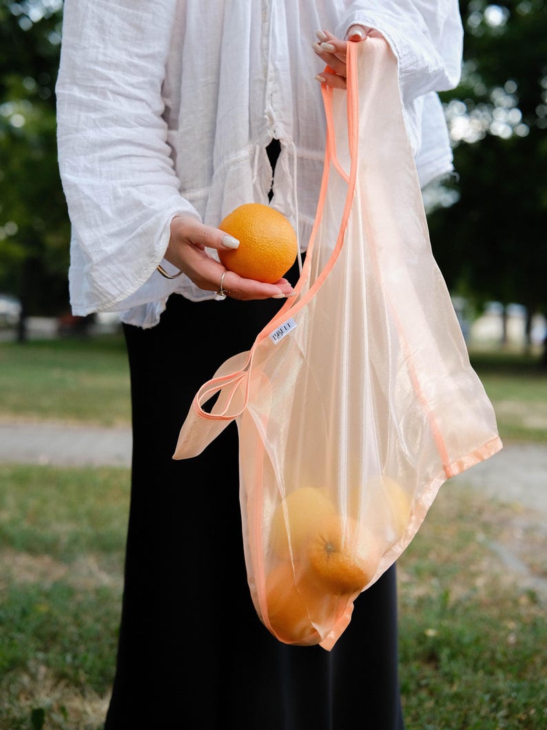 May include: A person holding a peach-colored mesh bag with oranges inside. The bag has a label that says "FREI."