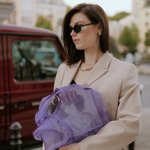 K&ouml;nnte beinhalten: Eine Frau in einem beige Blazer und Sonnenbrille h&auml;lt eine lilafarbene Netz-Tasche mit einem Magazin darin. Das Magazincover zeigt eine Frau mit lockigem Haar.