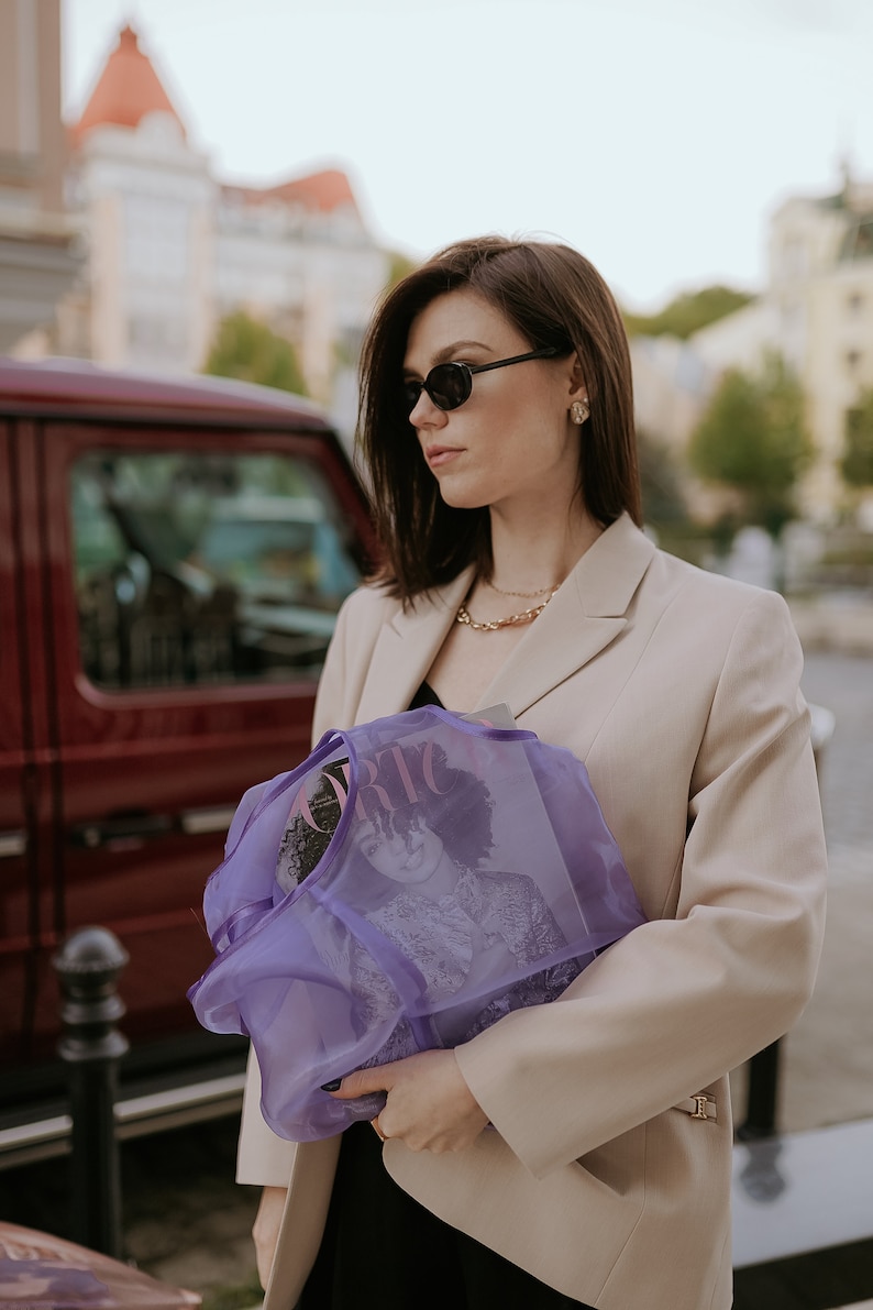 K&ouml;nnte beinhalten: Eine Frau in einem beige Blazer und Sonnenbrille h&auml;lt eine lilafarbene Netz-Tasche mit einem Magazin darin. Das Magazincover zeigt eine Frau mit lockigem Haar.