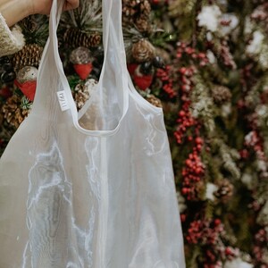 May include: A sheer white tote bag with long handles is held up against a backdrop of a Christmas tree. The bag has a tag with the text "1811" on it. The bag is made of a lightweight, translucent material.