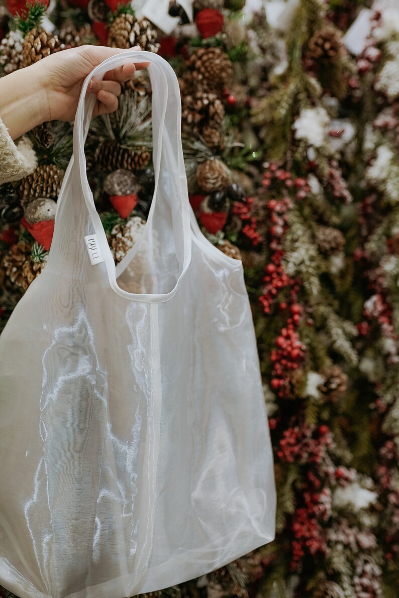 May include: A sheer white tote bag with long handles is held up against a backdrop of a Christmas tree. The bag has a tag with the text "1811" on it. The bag is made of a lightweight, translucent material.