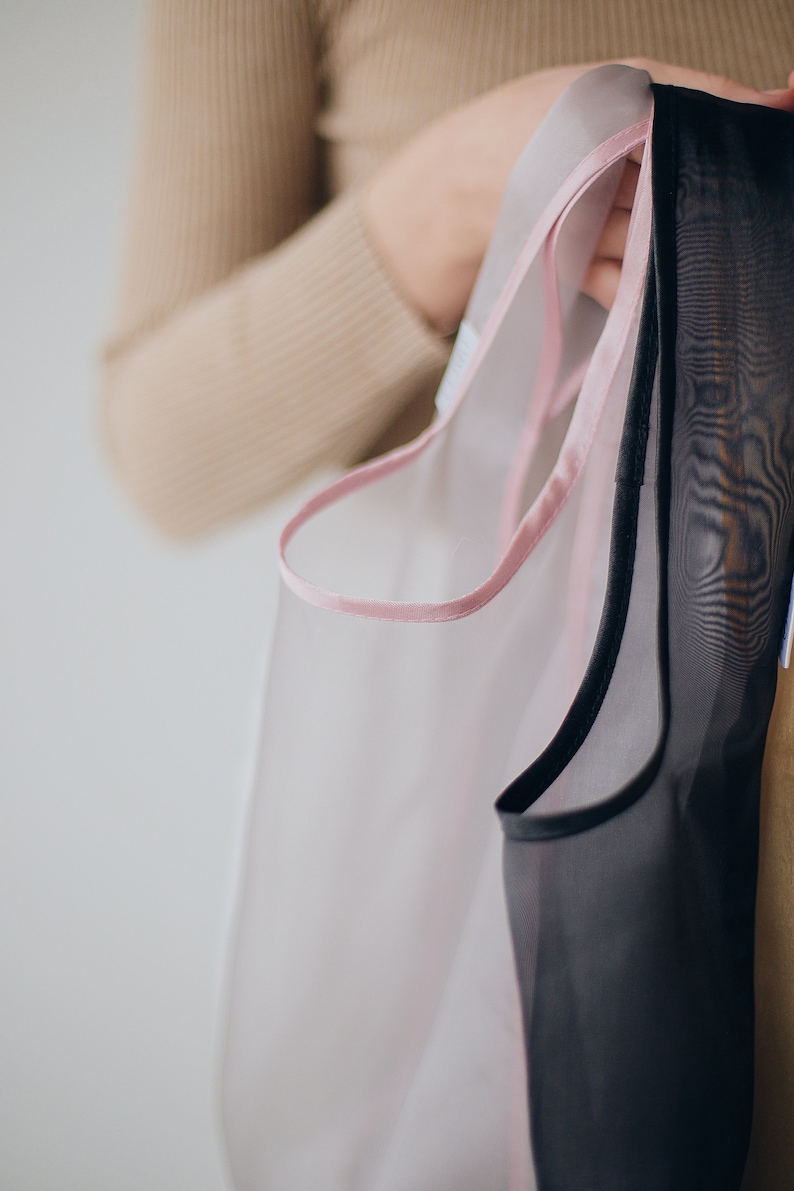 May include: Two sheer fabric bags with pink and black trim. The bags are held by a hand.