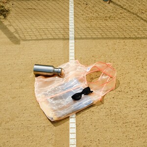 May include: A pink mesh tote bag with a silver water bottle and sunglasses inside, lying on a sandy surface with a white line.