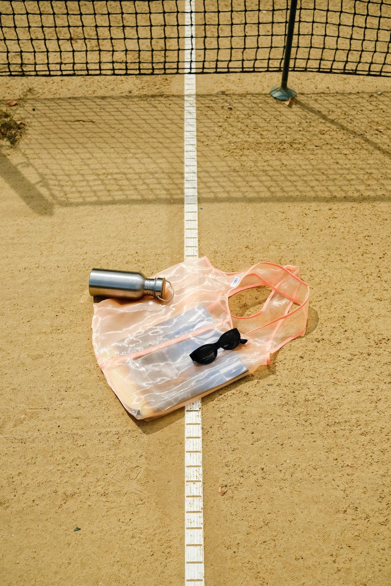 May include: A pink mesh tote bag with a silver water bottle and sunglasses inside, lying on a sandy surface with a white line.