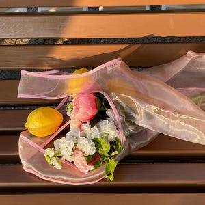May include: A transparent pink tote bag with a pink trim, filled with a bouquet of white and pink flowers, a pink peony, and a lemon. The bag is sitting on a brown wooden bench.