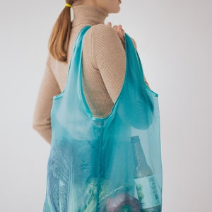 May include: A person carrying a turquoise mesh tote bag filled with groceries, including a bottle of beer, a head of cabbage, and a red bell pepper.