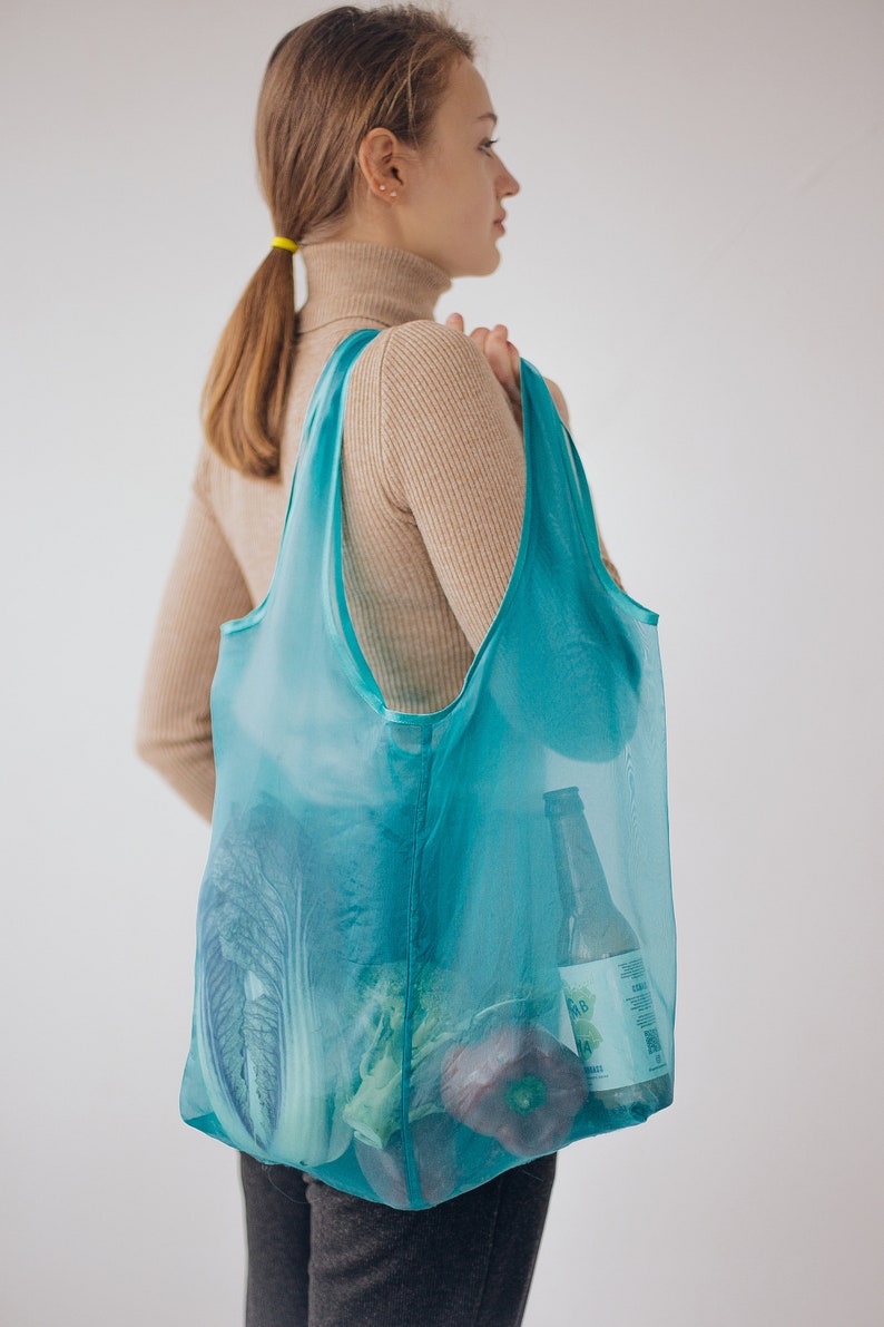 May include: A person carrying a turquoise mesh tote bag filled with groceries, including a bottle of beer, a head of cabbage, and a red bell pepper.