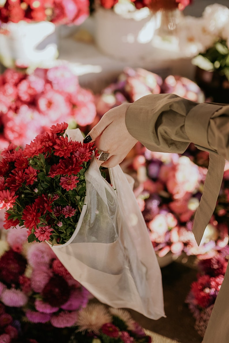 May include: A bouquet of deep red chrysanthemums in a white mesh bag, being held by a hand with a ring. The flowers are surrounded by a blur of pink and purple flowers.
