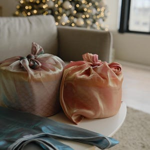 May include: Two gifts wrapped in pink and blue fabric with a decorative knot on top. The gifts are sitting on a white table with a blue fabric on top.