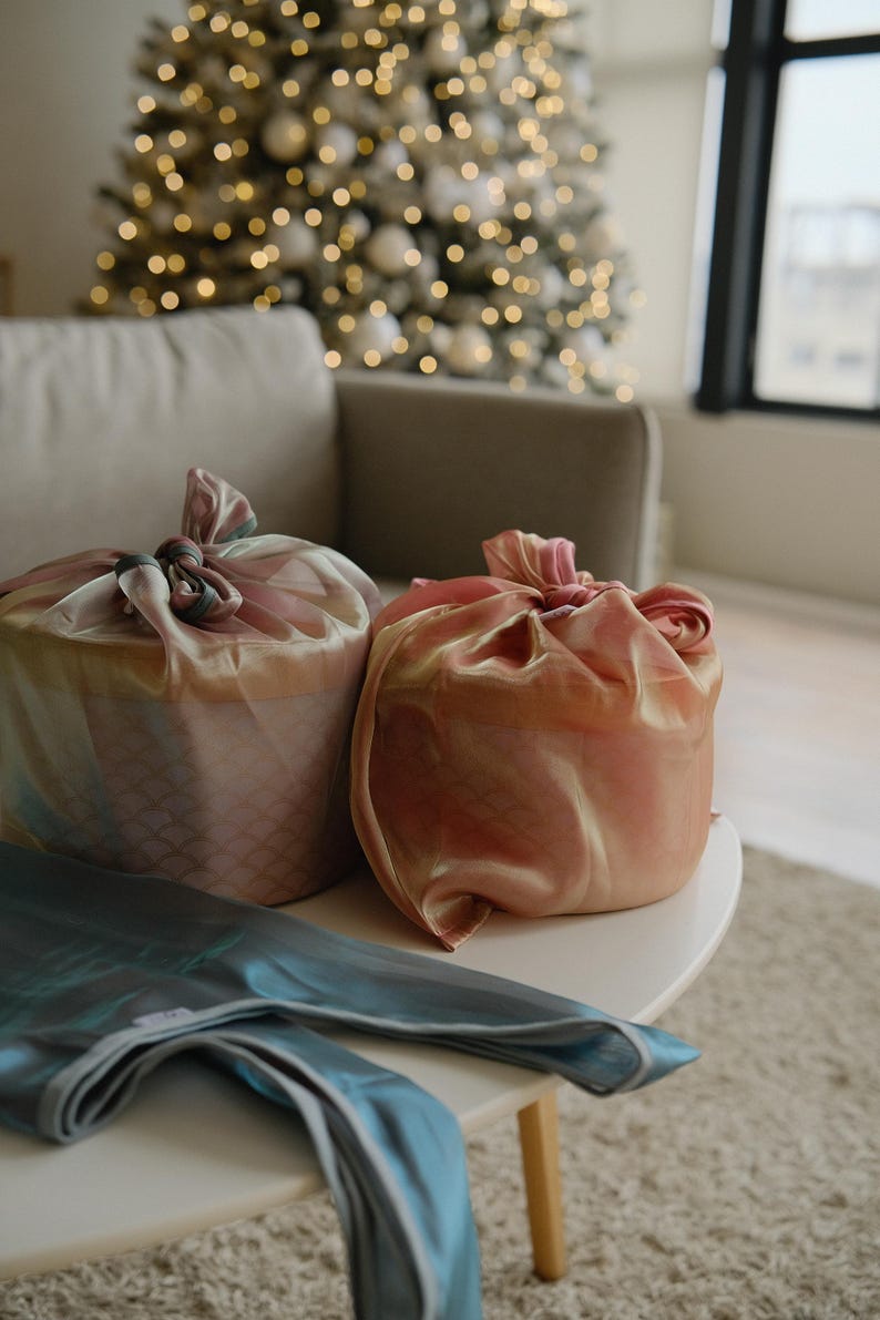 May include: Two gifts wrapped in pink and blue fabric with a decorative knot on top. The gifts are sitting on a white table with a blue fabric on top.