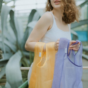 K&ouml;nnte beinhalten: Eine Frau h&auml;lt zwei wiederverwendbare Einkaufstaschen, eine gelbe und eine lila. Die Taschen sind aus einem durchsichtigen, leichten Material.