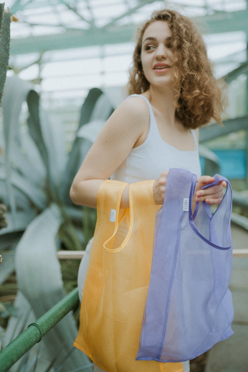 K&ouml;nnte beinhalten: Eine Frau h&auml;lt zwei wiederverwendbare Einkaufstaschen, eine gelbe und eine lila. Die Taschen sind aus einem durchsichtigen, leichten Material.