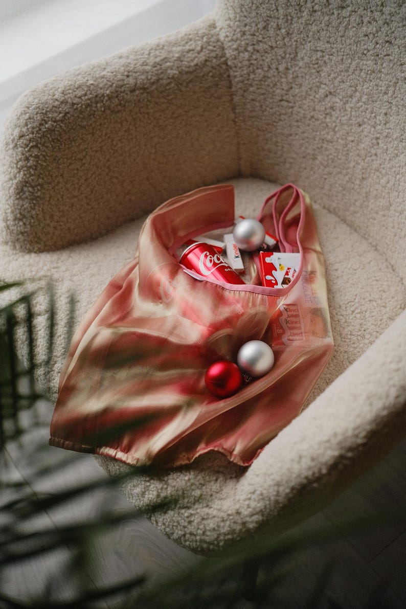 May include: A pink mesh tote bag filled with a can of Coca-Cola, red and silver baubles, and other small items. The bag is sitting in a fluffy white chair.