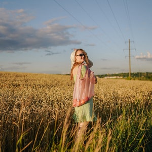 May include: A woman in a green dress and sunglasses stands in a field of wheat, holding a pink mesh bag.