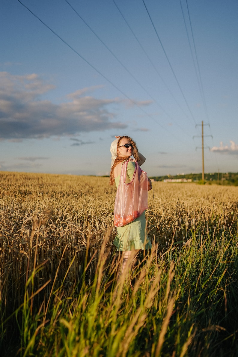 May include: A woman in a green dress and sunglasses stands in a field of wheat, holding a pink mesh bag.