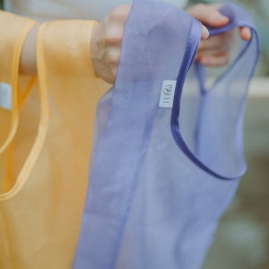K&ouml;nnte beinhalten: Eine Person h&auml;lt zwei wiederverwendbare Einkaufstaschen. Eine Tasche ist gelb und die andere ist lila mit dem Wort "LIBELLI" darauf gedruckt.