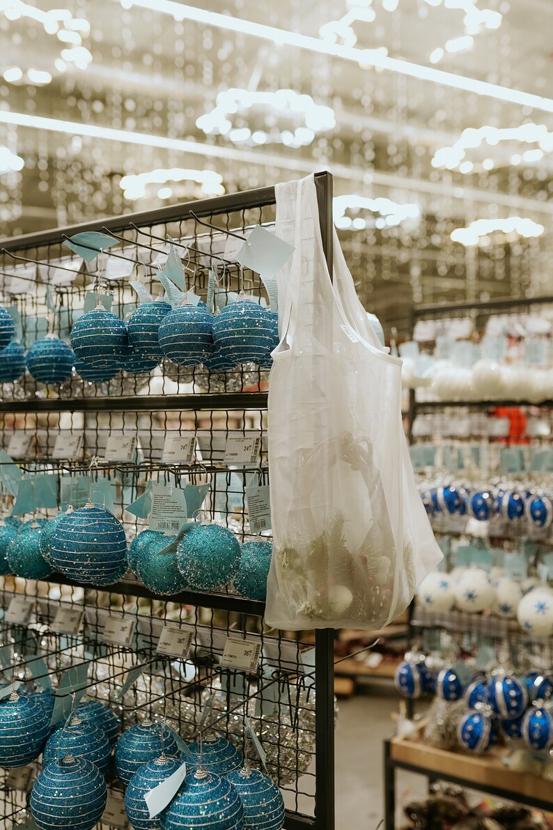 May include: A display of Christmas ornaments in a store. The ornaments are various shades of blue and white, with glitter and striped designs. A clear plastic bag hangs on the display, filled with more ornaments and greenery.