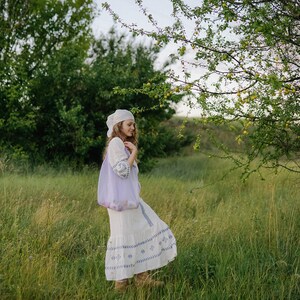 May include: A woman in a white dress with blue embroidery and a white headscarf walks through a field of green grass.