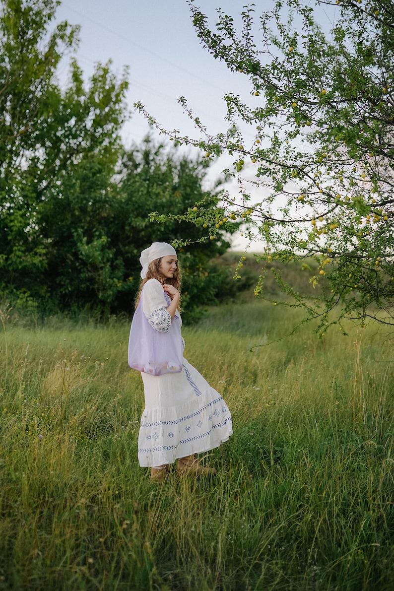 May include: A woman in a white dress with blue embroidery and a white headscarf walks through a field of green grass.