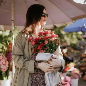 Puede incluir: Una mujer con gafas de sol y una gabardina beige sostiene un ramo de flores rosas y rojas envuelto en papel blanco.