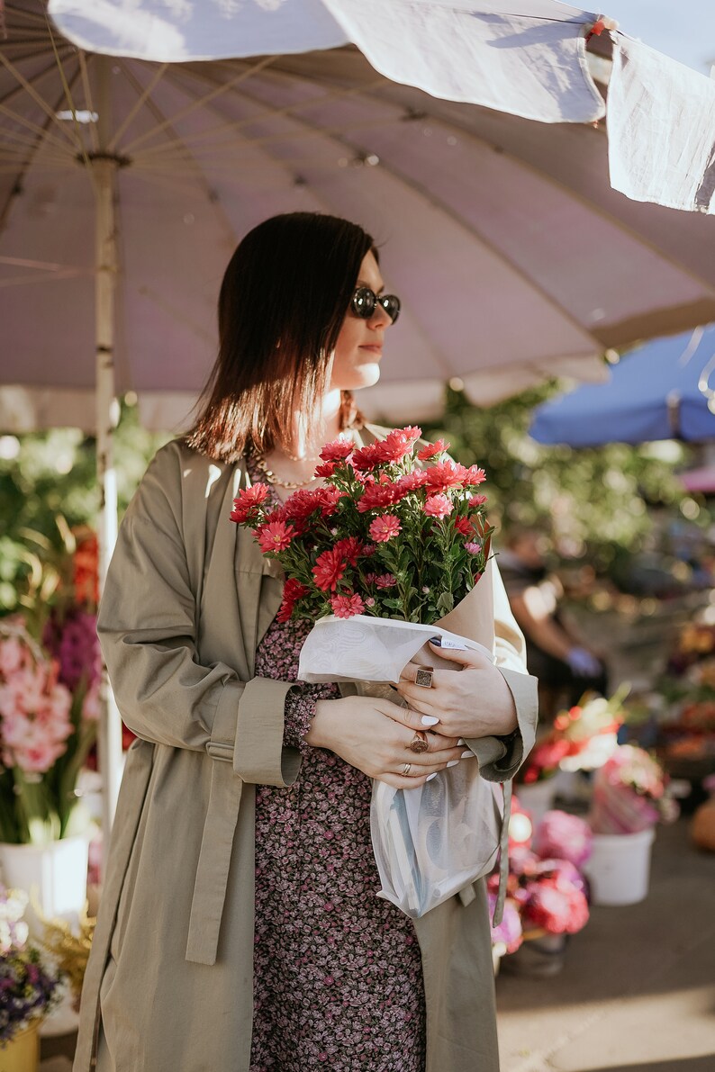 Puede incluir: Una mujer con gafas de sol y una gabardina beige sostiene un ramo de flores rosas y rojas envuelto en papel blanco.