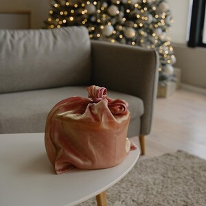 May include: A round gift box wrapped in a pink and gold fabric with a bow on top. The box is sitting on a white coffee table in front of a sofa and a Christmas tree.