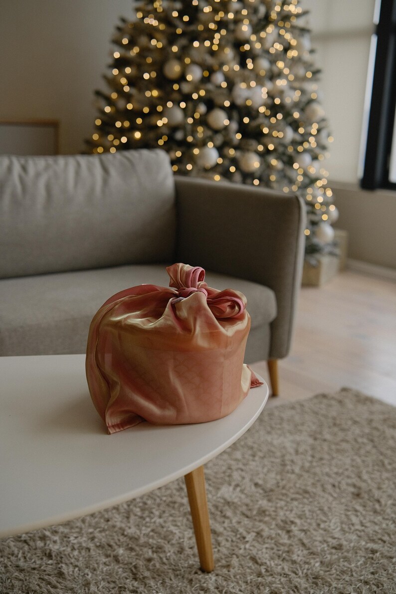 May include: A round gift box wrapped in a pink and gold fabric with a bow on top. The box is sitting on a white coffee table in front of a sofa and a Christmas tree.