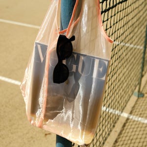 May include: A sheer pink mesh tote bag with a pink trim, holding a copy of Vogue magazine and a pair of black sunglasses. The bag is hanging on a blue metal pole.