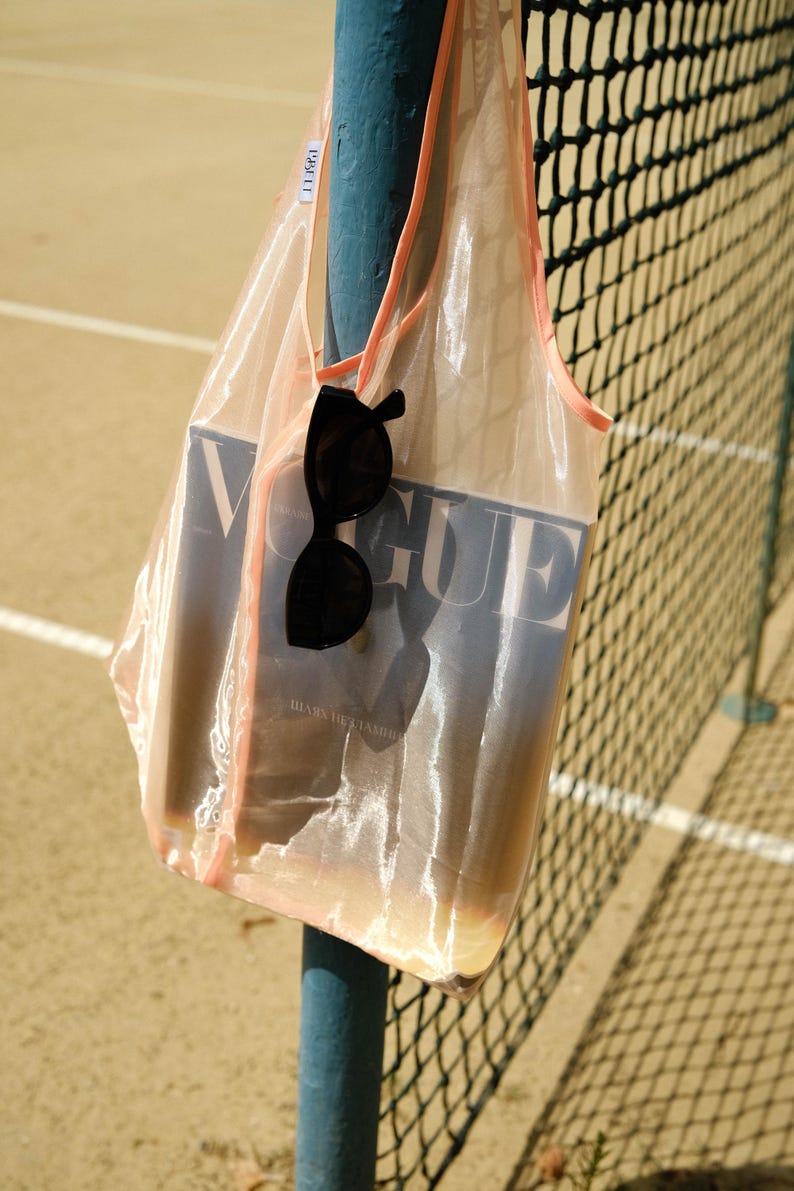 May include: A sheer pink mesh tote bag with a pink trim, holding a copy of Vogue magazine and a pair of black sunglasses. The bag is hanging on a blue metal pole.