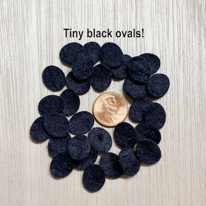 May include: A collection of small black felt ovals, scattered on a wooden surface. The ovals are about the size of a US penny.