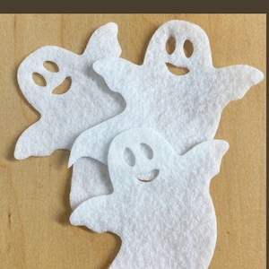 May include: Three white felt ghost shapes with smiling faces, cut out and arranged on a wood background.