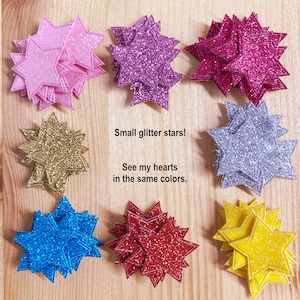 May include: A set of eight glitter star shapes in various colors, including pink, purple, red, gold, blue, silver, and yellow. The stars are stacked on top of each other and are arranged in a circle on a wooden surface. The text "Small glitter stars! See my hearts in the same colors." is written below the stars.