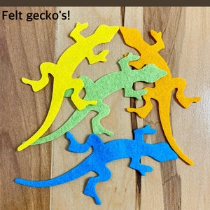 May include: Four felt gecko shapes in different colors: yellow, green, blue, and orange. The geckos are arranged in a playful, overlapping pattern. The text "Felt gecko's!" is visible in the top left corner of the image.