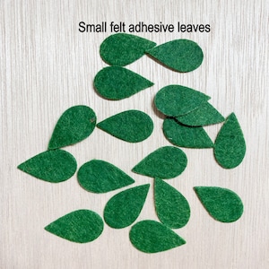 May include: A collection of small green felt leaves with adhesive backing. The leaves are shaped like teardrops and are scattered on a light wood surface.