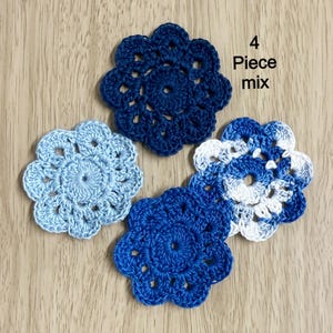 May include: Four crocheted flower appliques in shades of blue and white are arranged on a wooden surface. The appliques have a floral design with detailed stitching. The text "4 Piece mix" is visible in the image.
