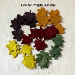 May include: A collection of tiny felt maple leaves in various fall colors, including yellow, orange, red, green, and dark purple. The text "Tiny felt maple leaf mix" is at the top of the image. These leaves are arranged in a circular pattern.