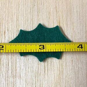 Może przedstawiać: Zielony filcowy liść ostrokrzewu o szerokości 7,6 cm.