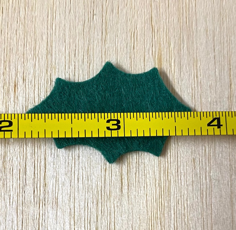 Może przedstawiać: Zielony filcowy liść ostrokrzewu o szerokości 7,6 cm.