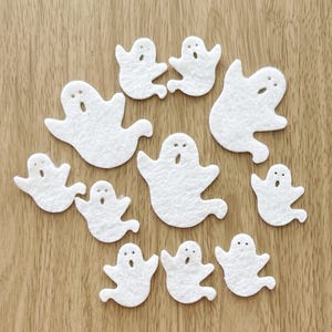 May include: A collection of eleven white felt ghost shapes with open mouths and dot eyes, arranged on a light brown wooden surface. The ghosts vary in size and are suitable for Halloween crafts and decorations.